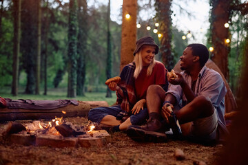 Happy couple of campers talking while eating hot dog by bonfire in nature.