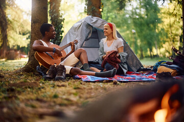 Young black camper playing acoustic guitar to his girlfriend in forest.
