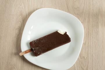 Creamy ice cream on a stick with dark chocolate on a white plate, top view. Natural light