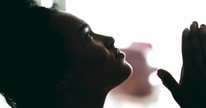 One spiritual young black woman looking up at sky in PRAYER feeling hopeful and faithful to a higher power. African American 20s person seeking God's support and help, profile close-up