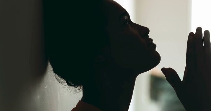 One spiritual young black woman PRAYING to GOD leaning on wall in profile silhouette. Person seeking help and support during difficult times with hands clenched looking up with HOPE
