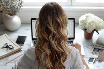 Rear view of a focused woman with curly hair working on a computer in a bright and organized office space with floral decor