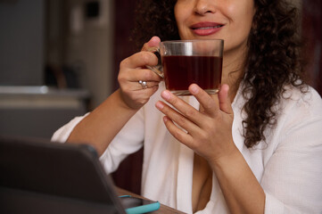 Details on hands of a blurred smiling pretty woman holding a cup of hot tea drink, sitting at table with digital tablet in modern home interior