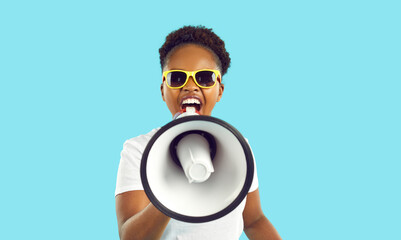 Cheerful crazy african american woman with loudspeaker in hand making loud advertisement on light blue background. Young woman in white t-shirt and sunglasses is shouting loudly about crazy discounts.