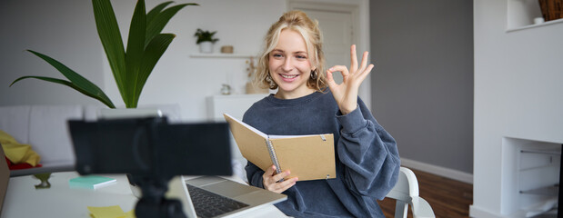 Portrait of smiling young woman, holding notebook, showing okay, ok sign, looking at digital...
