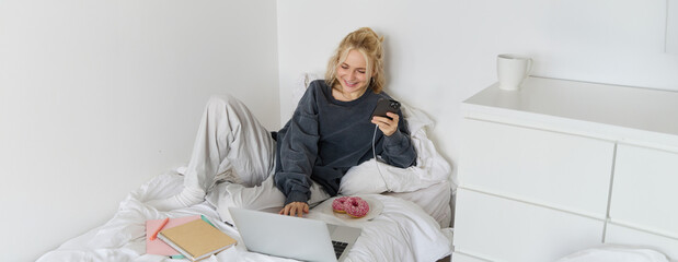 Portrait of smiling candid woman, lying in bed with doughnut, using smartphone and laptop, resting at home in bedroom, watching tv show or chatting online