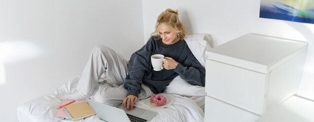 Portrait of young woman, student studying in her bed, relaxing while preparing homework, eating doughnut, using laptop in bedroom and drinking tea