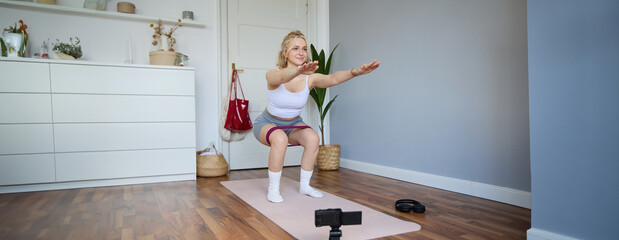 Portrait of woman, fitness instructor at home, recording video about workout, showing how to do leg...