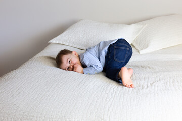 Adorable toddler boy lying down on bed in shirt and pants with happy and mischievous expression