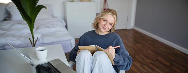 Portrait of smiling cute woman, lifestyle blogger, sits in her room with daily journal or planner,...