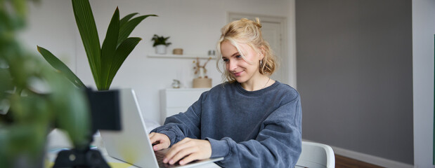 Portrait of young blond woman, female college student works from home on assignment, uses laptop, studies remotely, sits in room in front of computer