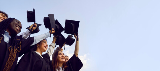 Group of happy multiethnic high school, college or university students having fun on graduation day and raising their graduate hats up to clear blue sky. Copy space banner background - 792007499