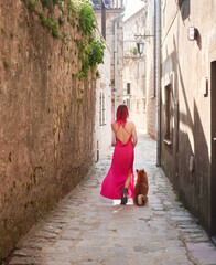 A woman in a striking pink dress strolls down an ancient cobblestone lane with her Shiba Inu dog,...