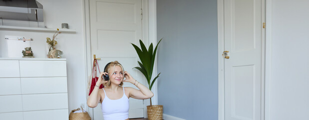 Portrait of cute young fitness woman, connects to online training session via laptop, wearing wireless headphones during workout, sits at home on rubber yoga mat