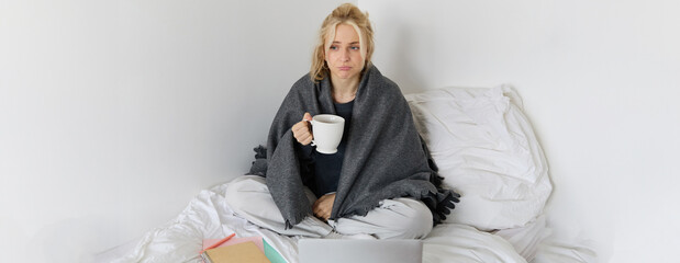 Portrait of woman catching a flu, sneezing, feeling sick, sitting on bed with laptop and working on...