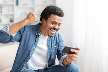 Cheerful black guy celebrating with smartphone at home