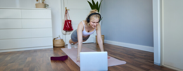 Portrait of young sporty woman doing workout, looking at fitness video on laptop in wireless headphones, standing in plank on yoga mat, following exercise tutorial
