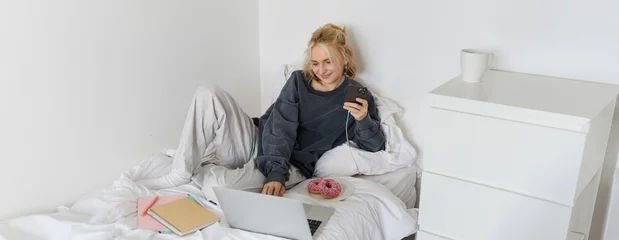 Foto op Plexiglas Lifestyle and weekend concept. Young smiling woman, relaxing at home, lying in bed with cup of tea and doughnut, using laptop and smartphone in bedroom © Mix and Match Studio