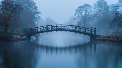 Fototapeta premium Foggy bridge in park at dawn. Mysterious and serene landscape photography. Mist, trees, and water reflection concept