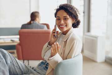 Smiling business woman sitting on office background and talking by phone with colleague