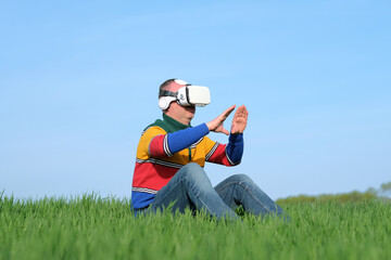 Amazed man touching air during the VR experience sitting on green grass in virtual reality glasses,...