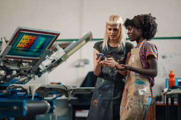 Interracial female graphic technicians at print shop looking at phone.