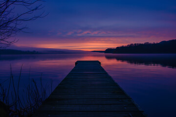 Obraz premium A wooden pier is seen in front of a body of water. The water is calm and the sky is a beautiful shade of purple. The scene is serene and peaceful, with the water reflecting the colors of the sky
