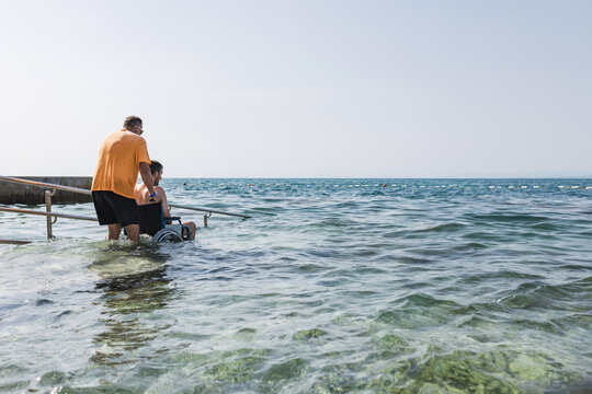 Male assistant helping a man in a wheelchair to enjoy the sea on an access ramp into the water. Accessible beach concept.