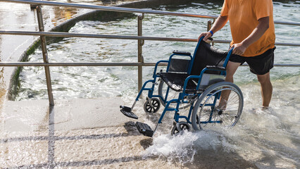Male assistant helping a man in a wheelchair to enjoy the sea on an access ramp into the water....