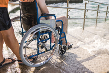 Male assistant helping a man in a wheelchair to enjoy the sea on an access ramp into the water....