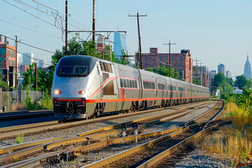 A train is traveling down the tracks in a city. The train is long and has many windows. The city is bustling with activity, and the train is a symbol of progress and modernity