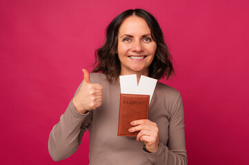 Studio portrait of a positive mid age woman holding passport and thumb up over pink background.
