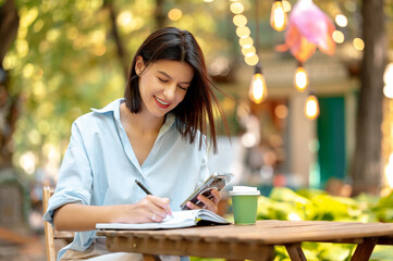 Young woman is using phone and enjoying coffee while writing in her planner in park on a sunny day.
