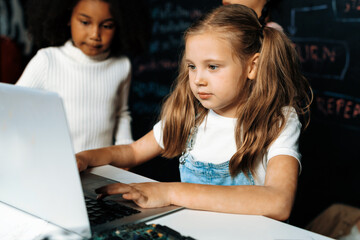 Blonde hair schoolgirl in white bib learning about coding robotics technology using laptop in the STEM class with confident look. White sweater schoolgirl look her coding with curious eyes. Erudition.