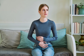 Young teenage female experiencing abdominal pain, sitting on couch at home