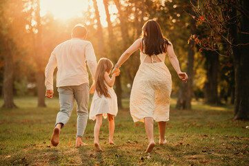 Naklejka na ściany i meble Young family of three are holding hands and running barefoot in park together at sunset.