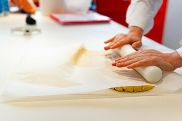 two hands knead yellow cookie dough with a rolling pin between baking papers on a kitchen table with a recipe notebook