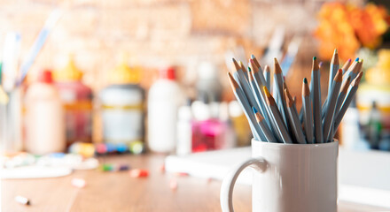 White mug with colored pencils in the foreground on a wooden table with art supplies in the...
