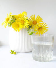 A bouquet of yellow flowers in a white vase and a glass glass with water on a white background. Postcard, place for text.