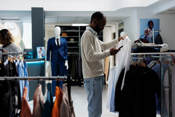 Retail market boutique customer examining hanging garment on rack while selecting size and style in...