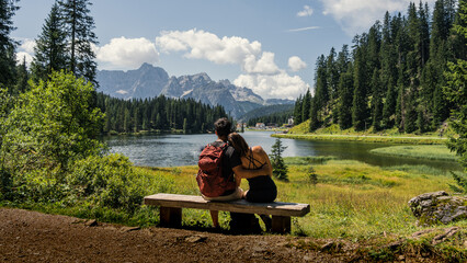 Couple sitting on a bench admiring views of a lake surrounded by mountains and pine trees