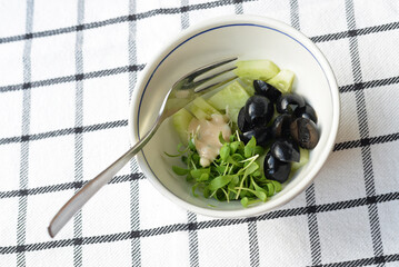Green and black salad with cucumber, watercress and black olives with a drop of cocktail sauce and a fork in a white bowl on a black and white checkered cloth