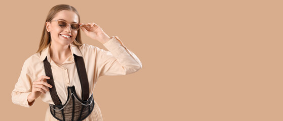 Stylish young woman in sunglasses with suspenders on beige background