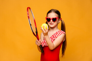 Cheerful little sports girl in red swimsuit playing beach tennis isolated over yellow background....