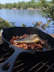 A smallmouth bass in a cast iron pan with bacon cooking over a fire beside the lake in the summer