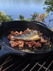 A smallmouth bass in a cast iron pan with bacon cooking over a fire beside the lake in the summer