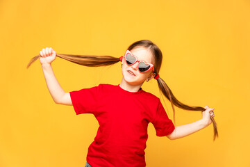 The happy little girl in red t-shirt and red summer sunglasses lifts her ponytails of hair up, and...