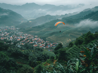 A paraglider flies over rolling hills and a quaint village shrouded in mist.
