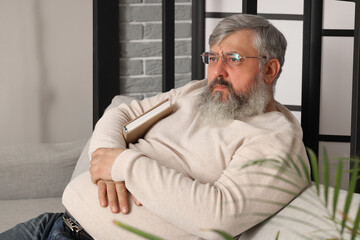Portrait of sad senior man with book sitting on sofa in living room - obrazy, fototapety, plakaty