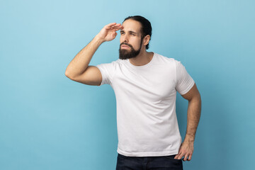 Side view of curious man with beard wearing T-shirt keeping palm over head and looking attentively...